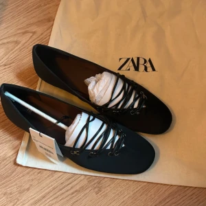 Svarta ballerinaskor med snörning från Zara - Svarta ballerinaskor från Zara med snygg snörning över foten och rund tå. Skorna har en platt sula och är tillverkade i ett syntetmaterial med Airfit technical insole för extra komfort. Perfekt för dig som gillar stilrena och trendiga detaljer.