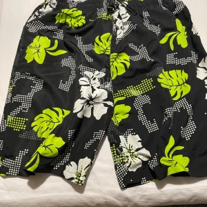 Svarta blommiga badshorts Runtech XL - Snygga svarta badshorts från Runtech i storlek XL med limegröna och vita blommor och blad. Shortsen har elastisk midja med snörning och meshfoder på insidan. Perfekta för stranden eller poolen, riktigt chill vibe och coolt mönster.