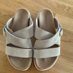 Beige mockasandaler från Sweeks - Snygga sandaler från Sweeks i beige mocka med breda korslagda remmar och en dekorativ spänndetalj i guld. Skorna har öppen tå och platt sula, perfekt för varma dagar. Stilren design som passar till många outfits. I ny skick använda en ända gång 