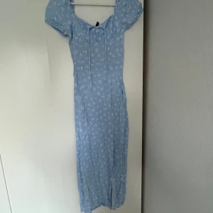 Ljusblå långklänning med blommor - Superfin ljusblå långklänning med vitt blommigt mönster och puffärmar. Klänningen har snörning vid halsen och en slits framtill som ger en snygg touch. 