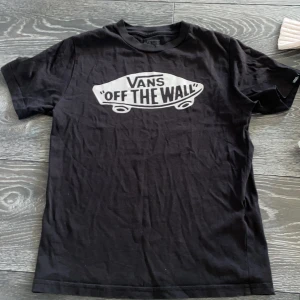 Svart Vans t-shirt med tryck - Svart t-shirt från Vans med klassisk 'Off The Wall'-logga i vitt framtill. Modellen har rund hals och korta ärmar, tillverkad i 100% bomull för skön och avslappnad känsla. Perfekt för dig som gillar streetwear och skate-vibe.