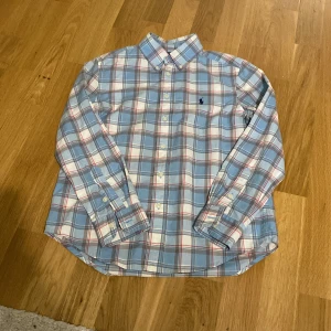 Rutig skjorta från Ralph Lauren - Snygg rutig skjorta från Ralph Lauren i ljusblått, vitt och röda detaljer. Klassisk krage, långärmad och med knappar framtill. Skjortan har det ikoniska Polo-logot broderat på bröstet. 