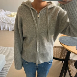 Grå hoodie med dragkedja från Zara - Mysig grå hoodie från Zara i storlek M. Tröjan har huva med snören, hel dragkedja framtill och breda ribbade muddar vid ärmslut och nederkant. Materialet är en blandning av akryl, polyamid och ull som gör den både mjuk och varm. 