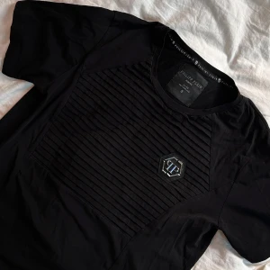 Svart Philipp Plein t-shirt med emblem - Svart t-shirt från Philipp Plein med ribbad panel framtill och ett metalliskt PP-emblem på bröstet. Rund hals och korta ärmar. Snyggt och stilrent plagg med coola detaljer, perfekt för dig som gillar exklusiva streetwear-vibbar.