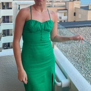 Grön långklänning med slits - Snygg grön långklänning med smala axelband som korsas i ryggen och knyts. Klänningen har en slits framtill, knappar vid bysten och smock i ryggen för skön passform. Perfekt för sommarkvällar och har en feminin, figurnära siluett.