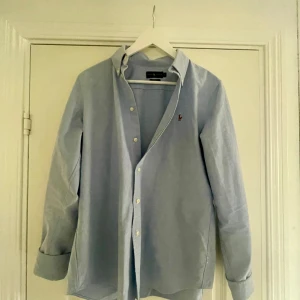 Ljusblå skjorta från Ralph Lauren - Snygg ljusblå skjorta från Ralph Lauren med klassisk krage och knappar framtill. Liten broderad logga i rött på bröstet. Skjortan har lång ärm och är gjord i mjuk bomull, perfekt för en clean och chill stil.nu pris 1600kr säljer endast för 499kr