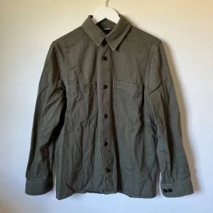 Olivgrön overshirt från ARKET - Olivgrön overshirt från ARKET i ekologisk bomullstwill. Skjortan har två stora bröstfickor, klassisk krage och svarta knappar framtill samt vid ärmslut. Regular fit och långärmad, perfekt för lager-på-lager. Snygg och enkel att matcha med jeans eller chinos. 1A19
