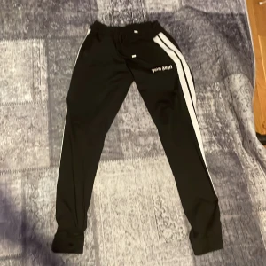 Palm Angels svarta mjukisbyxor - Svarta mjukisbyxor från Palm Angels med vita sidoränder och logotyp på höften. Byxorna har snörning i midjan och är gjorda i ett mjukt syntetmaterial. Passformen är slim och de har muddar vid bensluten för en sportig vibe.