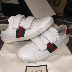 Gucci vita sneakers  - Säljer ett par vita Gucci sneakers med klassiska gröna och röda ränder på sidan. Skorna har två breda kardborreband och detaljer i metallicrött och metallicgrönt på hälen. Tillverkade i skinn och har rund tå samt platt sula. Perfekt för dig som gillar lyxiga och stilrena sneakers. Kommer med låda och skopåse 