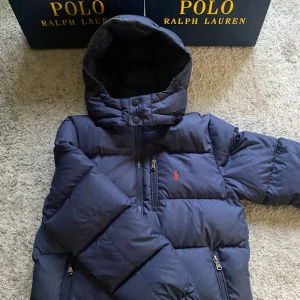 Mörkblå pufferjacka från Polo Ralph Lauren - Snygg mörkblå pufferjacka från Polo Ralph Lauren med huva och flera dragkedjor. Jackan har ett klassiskt rött Polo-logo på bröstet och är vadderad för extra värme. Perfekt för kalla dagar och har en normal passform med långa ärmar.