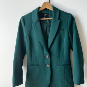 Grön kavaj från H&M, storlek 36 - Snygg grön kavaj från H&M i storlek 36. Kavajen har två knappar framtill, klassisk krage och figurnära passform. Fickor med diskreta öppningar och helfodrad insida. Perfekt för dig som gillar stilrena och moderna plagg.