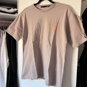 Beige t-shirt med tryck - Säljer en beige t-shirt från Lager157 med ett coolt orange tryck av en smiley med blixt och stjärna. T-shirten är kortärmad och gjord i mjuk bomull. Perfekt för en avslappnad stil. Önskar du veta exakta mått? Tveka inte att höra av dig!