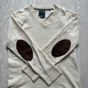 Beige tröja från Marc O'Polo - Säljer en stilren beige tröja från Marc O'Polo med v-ringning och bruna armbågslappar. Tröjan har långa ärmar och ribbade kanter vid hals och ärmslut. Perfekt för en avslappnad och elegant look.