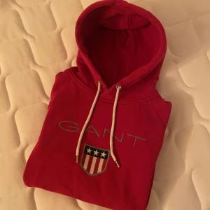 Röd hoodie från Gant - Säljer en snygg röd hoodie från Gant med en stor logga och ett sköldmärke på framsidan. Den har en justerbar huva med vita snören och är perfekt för en avslappnad stil.