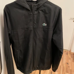 Lacoste jacka - Snygg svart vindjacka från Lacoste med dragkedja och huva. Jackan har en broderad krokodil-logga på bröstet och elastiska muddar vid ärmslut och nederkant. Perfekt för blåsiga dagar! Säljer då den har blivit för liten