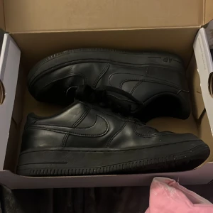 Svarta Nike Air Force sneakers - Säljer ett par klassiska svarta Nike Air Force sneakers. Skorna har en stilren design med snörning och är tillverkade i syntetmaterial. Perfekta för en casual look.