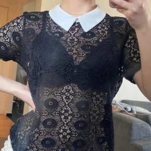 Svart spetsblus med vit krage - Snygg svart spetsblus med en kontrasterande vit krage. Blusen har korta ärmar och ett vackert cirkulärt mönster i spetsen. Perfekt för att ge en elegant touch till din outfit.