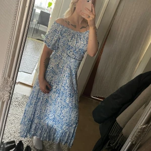 Blå och vit långklänning - Blå/vit paisley mönstrad långklänning i mjukt och följsamt material. Går även att bära som off-shoulder. Volang nedtill och resår i ärm och midja. 100% mjuk viskos. Strl M 