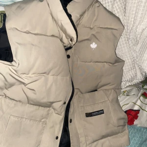 Beige dunväst från Canada Goose - Säljer en snygg beige dunväst från Canada Goose med broderad lönnlöv på bröstet. Västen har dragkedja och praktiska fickor framtill. Perfekt för kyligare dagar.