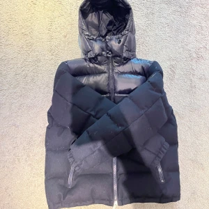 Mörkblå dunjacka från Polo Ralph Lauren - Snygg svart dunjacka från Polo Ralph Lauren med huva och dragkedja. Jackan har en quiltad design och två praktiska fickor med dragkedja framtill. Perfekt för kyliga dagar när du vill hålla stilen. 