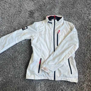 Vit vindjacka från Helly Hansen - Säljer en vit vindjacka från Helly Hansen med dragkedja och hög krage. Jackan har lite fläckar och är i bra skick men använd