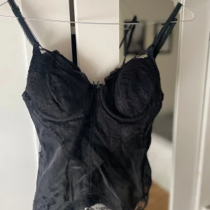 Svart spetskorsett - Elegant svart korsett med spetsdetaljer och justerbara axelband. Perfekt för en stilfull look med en feminin touch. Korsetten har en figurnära passform och vacker spetskant nedtill.