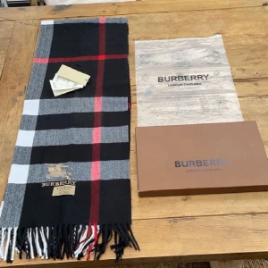 Burberry Halsduk - Säljer nu denna super stilrena Burberry halsduken! Det är en mycket fin halsduk i Kashmir, fin färgkombination av grå, vit, mörkblå/svart och en touch av röd! Cond:10/10 finns inget som helst tecken av användning! Låda, plast, tags och halsduk kommer med!Kom Pm för minsta fråga! 🍾🥃