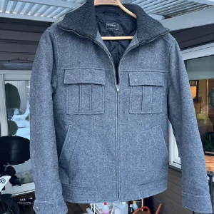 Grå ulljacka  - Snygg grå ulljacka  med dragkedja och ribbad krage. Jackan har två bröstfickor med lock och två sidofickor. Perfekt för kyligare dagar med sin quiltade insida.