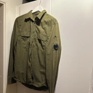 Grön overshirt från C.P. Company - Snygg grön overshirt från C.P. Company med klassisk krage och knappar framtill. Den har långa ärmar och en cool detalj med en lins på ärmen. Perfekt för lager-på-lager-stil. Storlek L 