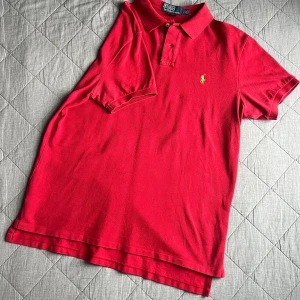 Röd pikétröja från Ralph Lauren - Säljer en klassisk röd pikétröja från Ralph Lauren med den ikoniska broderade loggan i grönt på bröstet. Tröjan har korta ärmar och en stilren krage med knappar. Perfekt för en avslappnad men ändå stilren look. Inga skavanker.