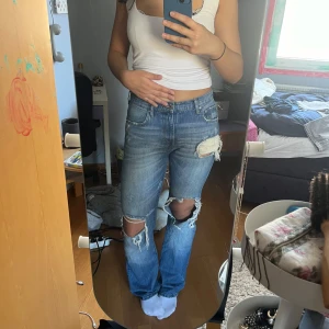 Blå jeans med slitningar från Zara - Säljer ett par blå jeans från Zara med coola slitningar på knäna och bakfickan. De har en dragkedja och knapp framtill. Perfekta för en avslappnad look.