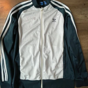 Vit och mörkgrön Adidas track jacket - Säljer en klassisk vit och mörkgrön Adidas track jacket med dragkedja. Nopprig o en fläck på framsidan. Se bilderna ovan