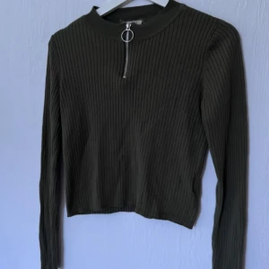 Mörkgrön ribbad tröja med dragkedja - Snygg mörkgrön ribbad tröja med dragkedja framtill. Tröjan har långa ärmar och en croppad passform, perfekt för en trendig look. Passar bra till både jeans och kjol.