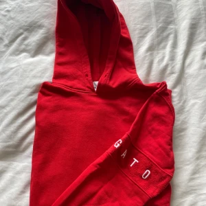 Axel Arigato hoodie - Säljer en snygg röd hoodie från Axel Arigato. Den har en klassisk design med en stor huva och textdetalj på ärmen. Perfekt för en avslappnad stil.