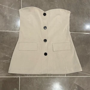 Beige axelbandslös klänning från Gina Tricot - Snygg beige axelbandslös klänning från Gina Tricot med knappar framtill och två fickor. Perfekt för en stilren look. Klänningen har en figurnära passform och är tillverkad i ett mjukt material.