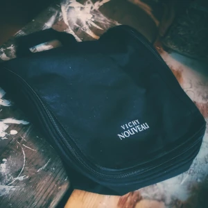 Svart necessär från Vichy Nouveau - Praktisk svart necessär från Vichy Nouveau med dragkedja och flera fack för förvaring. Perfekt för att organisera dina tillhörigheter. Den har en krok för enkel upphängning.