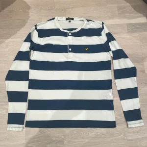 Randig långärmad tröja från Lyle & Scott - Säljer en stilren randig tröja från Lyle & Scott i blått och vitt. Tröjan har långa ärmar och knappar vid halsen. Den är dekorerad med Lyle & Scotts ikoniska logga på bröstet. Perfekt för en avslappnad stil. Tröjan är vit och blå randig. Det står att det är storlek L men det är i barn storlek så skulle säga S