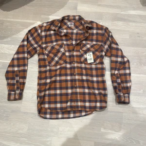 Rutig skjorta från Levi's - Snygg rutig flanellskjorta från Levi's i orange, blå och vit. Skjortan har långa ärmar och knappar framtill samt två bröstfickor. Perfekt för en avslappnad stil. Helt ny med lappen kvar. 