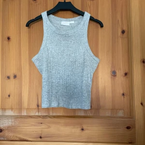 Grå ribbad linnetopp från Gina Tricot - Säljer en stilren grå ribbad linnetopp från Gina Tricot. Perfekt för en avslappnad look. Toppen har en klassisk rund halsringning och ärmlös design.