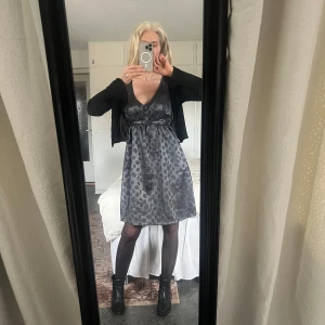 Grå prickig klänning från Soaked in Luxury - Snygg grå klänning med prickigt mönster från Soaked in Luxury. Klänningen har en v-ringad design och knappar framtill. Den är knälång och har ett knytband i midjan för en smickrande passform. Perfekt för en stilren look!