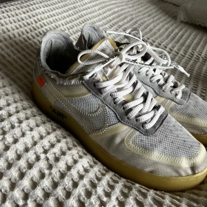 Nike Air Force sneakers i vitt och grått - Säljer ett par Nike Air Force sneakers i vitt och grått med genomskinliga detaljer och text på sidan. Skorna har en gulaktig sula och klassisk snörning. Perfekta för den som vill ha en unik stil.