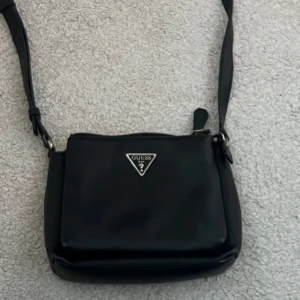 Svart axelväska från Guess - Snygg svart axelväska från Guess med justerbar axelrem och en stilren design. Väskan har en metallogo på framsidan och är perfekt för att bära dina nödvändigheter. Enkel att matcha med olika outfits.