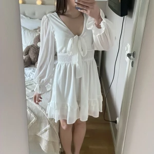 Super fin klänning - Säljer en söt vit klänning från Wodrow med v-ringning och knytband framtill. Klänningen har långa ärmar och en smockdetalj i midjan för en fin passform. Den har volanger vid fållen som ger en romantisk touch. Perfekt för sommarens alla tillfällen!