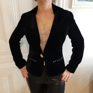 Svart kavaj med gulddetaljer - Snygg svart kavaj med guldfärgad knapp och dekorativa detaljer vid midjan. Perfekt för en stilren look. Kavajen har en klassisk passform och långa ärmar.