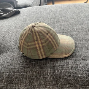 Rutig keps från Burberry - Snygg rutig keps från Burberry i gröna och bruna toner. Kepsen har en justerbar rem baktill med metallspänne för perfekt passform. Perfekt för en stilren look. Storlek L