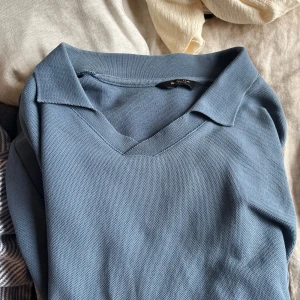 Blå pikétröja från Massimo Dutti - Snygg blå pikétröja från Massimo Dutti med klassisk krage och v-ringning. Perfekt för en stilren och avslappnad look. Tröjan har korta ärmar och är tillverkad i ett mjukt material som ger en bekväm passform.