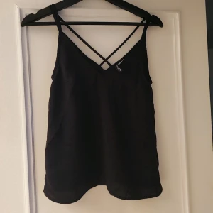 Svart linnetopp med korsade band - Snygg svart linnetopp med smala axelband och korsade band i ryggen. Perfekt för en stilren look. Toppen har en enkel design som gör den lätt att matcha med olika outfits.