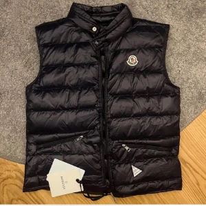 marinblå dunväst från Moncler - Snygg svart dunväst från Moncler med dragkedja och hög krage. Västen har två praktiska fickor med dragkedja och Monclers logga på bröstet. Perfekt för kyligare dagar.