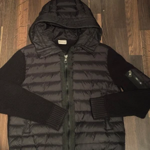 Svart Moncler cardigan - Snygg svart pufferjacka från Moncler med huva och dragkedja. Jackan har långa stickade ärmar och en quiltad kropp. Perfekt för kyligare dagar.