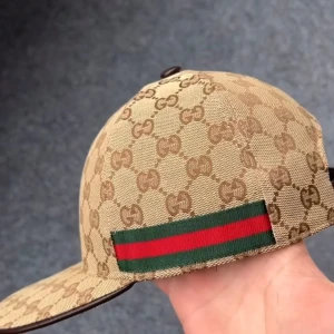 Beige keps från Gucci - Snygg beige keps från Gucci med det ikoniska GG-mönstret. Kepsen har en justerbar rem i brunt läder baktill och en grön och röd randdetalj på sidan. Perfekt för att ge en lyxig touch till din outfit. Har flera olika storlekar S,M,L